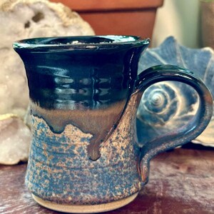 May include: A ceramic mug featuring a glossy dark navy blue glaze on the upper portion that drips down over a textured, speckled blue and tan base. The mug has a curved handle and a wide, flared rim, showcasing a handcrafted, multi-toned finish.