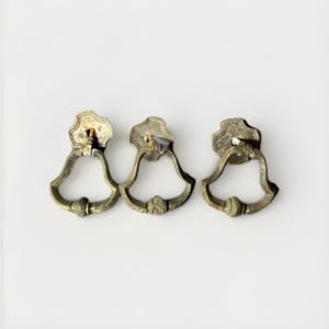 May include: Three antique-style brass drawer pulls featuring a bell-shaped drop handle attached to a decorative backplate. Each piece displays a weathered, tarnished metallic finish with a central knob detail on the bottom of the swinging loop.