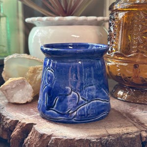 May include: A small, cylindrical ceramic vessel with a glossy, deep blue glaze. The exterior features a dark, incised line drawing of a reclining figure that wraps around the body of the piece. The rim is slightly flared and smooth in texture.
