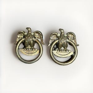 May include: A pair of antique-style brass drawer pulls, each featuring a detailed eagle perched atop a circular ring. The metal has a weathered, muted gold finish with visible texture on the wings and the semi-circular base beneath the eagle.
