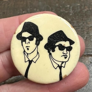 May include: A circular pinback button with an off-white background featuring a black line art illustration of two figures wearing fedoras, sunglasses, collared shirts, and neckties. The design depicts two individuals with similar attire.