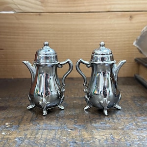 May include: A pair of silver-toned metal salt and pepper shakers shaped like miniature coffee pots. Each shaker features a curved handle, a small spout, a domed lid with perforated holes, and three decorative feet at the base of the body.