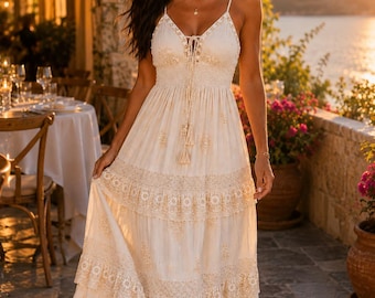 May include: A floor-length cream-colored maxi dress featuring thin spaghetti straps, a V-neckline with a lace-up tassel detail, and a smocked bodice. The skirt is designed with multiple tiered layers of fabric, accented by intricate embroidery.
