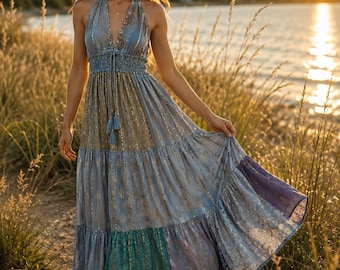 May include: A floor-length halter dress featuring a smocked waist with a drawstring and tassel detail. The dress displays a tiered skirt with a blue and gold metallic pattern, incorporating panels of teal and purple fabric at the lower hem.