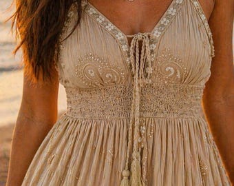 May include: A beige sleeveless dress featuring a V-neckline with white floral embroidery, a smocked elastic waist, and a central drawstring with two hanging tassels. The fabric has a textured, crinkled appearance with scattered white embroidery.