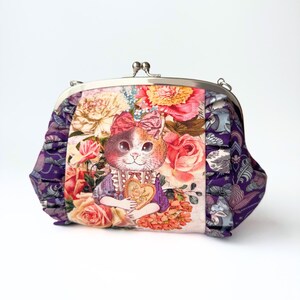 以下が含まれることがあります： 金属製の留め金が付いたヴィンテージスタイルのハンドバッグ。前面には、バラに囲まれたハートを持つ猫のイラストが描かれています。側面は紫と青の花柄で、気まぐれでロマンチックな美学を作り出しています。