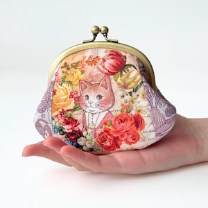 以下が含まれることがあります： 猫のイラストが入ったフローラルな小銭入れ。小銭入れは金属製の留め金が付いており、バラ、チューリップ、その他の花々のカラフルな模様で飾られています。猫はピンク色の服を着ています。