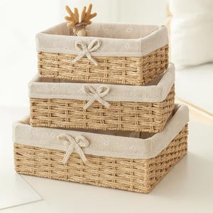 Peut inclure: Ensemble de trois paniers de rangement tressés de différentes tailles. Chaque panier est doublé d'un tissu beige clair et orné d'un nœud décoratif. Une petite figurine de cerf est posée dans le panier supérieur. Les paniers sont en matériau tressé de couleur naturelle.