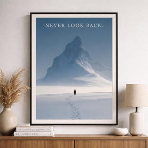 Pode incluir: Impressão artística emoldurada com uma paisagem montanhosa nevada sob um céu azul. A impressão inclui o texto "NEVER LOOK BACK". Uma figura solitária caminha pela neve, deixando pegadas. A obra de arte é exibida em um armário de madeira.
