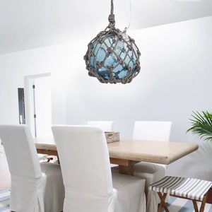 Puede incluir: Una lámpara colgante de globo de vidrio azul con un diseño de red de cuerda cuelga sobre una mesa de comedor con sillas blancas. La mesa es de madera y tiene un acabado marrón claro.