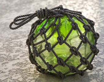 May include: A green glass buoy with a brown rope net. The buoy is round and has a smooth surface. The rope net is tightly woven and has a knot at the top.
