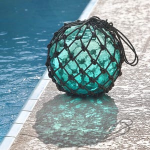 May include: A round, turquoise glass float with a black net covering sits on a concrete surface next to a pool. The float's shadow is visible on the concrete. The water in the pool is a deep blue.