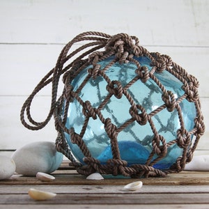 May include: A decorative blue glass float, encased in a brown rope netting, with a loop for hanging. The float is surrounded by white stones and seashells, evoking a nautical theme. The glass has a slight blue tint.