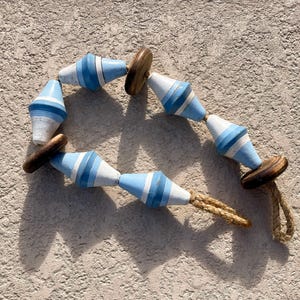 May include: A decorative garland featuring six diamond-shaped wooden beads painted in alternating stripes of light blue, white, and dark blue. The beads are interspersed with three dark brown circular wooden discs and strung on a thick jute rope.