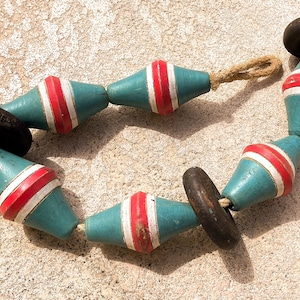 May include: A string of wooden buoys in a circle. The buoys are teal with red and white stripes. The end pieces are black and round. The buoys are connected by a natural rope.