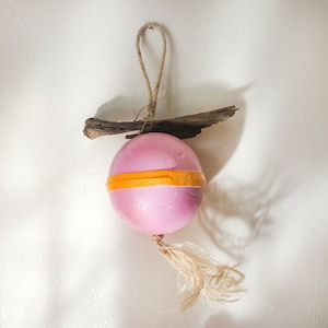 May include: A pink sphere ornament with an orange stripe, a natural wood accent, and a jute rope loop for hanging. A tassel hangs from the bottom. The ornament is a decorative item.