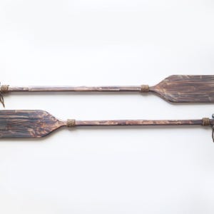 Op de afbeelding: Twee houten roeispanen met touwaccenten, hangend tegen een witte achtergrond.