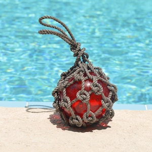 May include: A spherical red glass float encased in a decorative brown rope netting. The netting features a series of interconnected knots and extends upward to form a loop at the top for hanging. The glass surface is smooth and translucent.