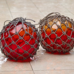 May include: Two red and orange glass floats with a gray rope net. The floats are round and have a textured surface.