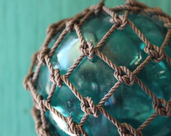 May include: A clear, teal-colored glass ball is encased in a knotted, brown rope netting. The nautical-themed decorative piece is suspended from a matching rope, creating a rustic, coastal aesthetic.