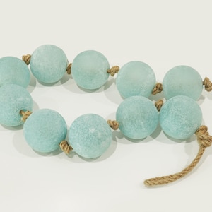 May include: A string of nine spherical, light blue, frosted glass floats connected by a thick, tan-colored jute rope. Each glass sphere features a textured, matte surface and is secured in place along the rope with small, hand-tied knots.