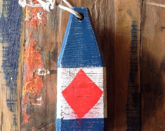 May include: A blue and white wooden buoy with a red diamond shape in the center, hanging by a white rope. The buoy has a distressed, weathered appearance, suggesting a nautical theme.