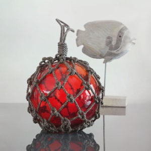 以下が含まれることがあります： 灰色のロープネットに包まれた赤いガラスフロート。フロートは丸く、質感のある表面をしています。