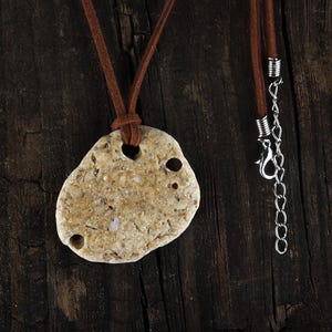 May include: A natural stone pendant necklace with a brown leather cord. The pendant is a light tan color with several holes and a unique, organic shape. The necklace has a silver-colored chain and clasp.