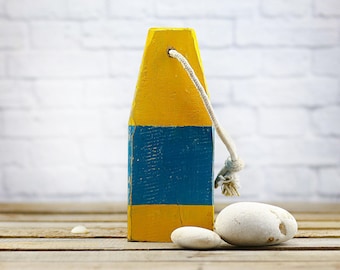 May include: A wooden buoy painted yellow and blue, with a white rope attached. Two smooth, white stones rest on a wooden surface in front of the buoy. The background is a white brick wall.