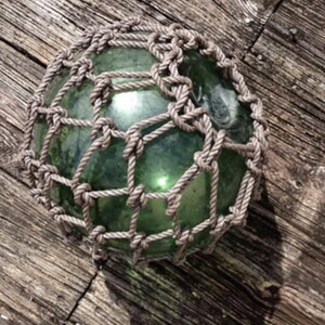 May include: A round, green glass float encased in a knotted rope net. The float is resting on weathered, wooden planks. The rope is a light brown color, and the glass has a slight green tint. The overall aesthetic is nautical.
