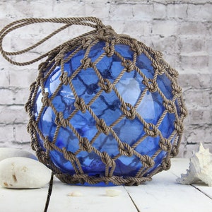 May include: A blue glass buoy, encased in a brown rope net, with a loop for hanging. The buoy is displayed with white stones and a seashell, evoking a nautical theme. The background is a white brick wall.