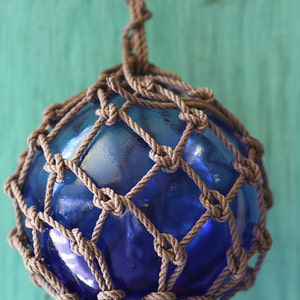 Puede incluir: Una boya de vidrio azul con una red de cuerda marrón. La boya está colgada de una cuerda.