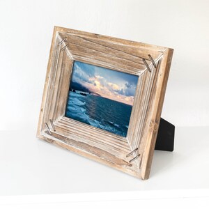 Puede incluir: Un marco de fotos de madera rústico con un acabado blanco desgastado. El marco tiene un caballete negro y presenta una fotografía de paisaje de una escena de playa con un cielo nublado.