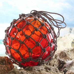 May include: A red glass buoy with a brown rope net. The buoy is sitting on a rocky surface with a beach and ocean in the background.