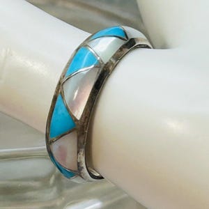 May include: A silver band ring featuring an inlay pattern of triangular and geometric segments. The inlay consists of alternating pieces of bright turquoise blue stone and iridescent white mother-of-pearl, set within a polished silver metal frame.
