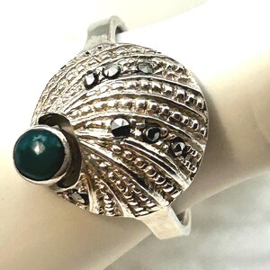 May include: A silver ring featuring a large, circular, textured face with a fan-like pattern. The ring is accented with a single, smooth, dark teal cabochon stone on the left side and several small, dark, faceted stones embedded across the surface.