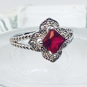 May include: A silver ring with a square, deep red gemstone at the center. The gemstone is surrounded by a decorative, textured setting. The band has a twisted rope design.