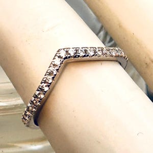 May include: A silver-toned, V-shaped ring, encrusted with small, sparkling gemstones. The ring's design features a row of stones along the top and sides, creating a continuous band of brilliance. The ring is set against a neutral background.
