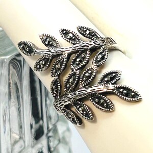 May include: A silver-toned ring featuring two overlapping branches with textured stems and leaves. Each leaf is inlaid with small, dark, faceted marcasite stones that provide a metallic shimmer. The design is open-ended, creating a bypass style.