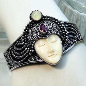 May include: A silver cuff bracelet featuring an intricate filigree design with a central carved cream-colored face with closed eyes. The piece is accented by a round iridescent opal at the top and an oval deep red gemstone set above the face.
