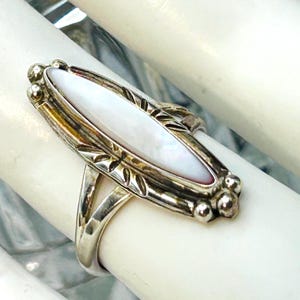 May include: A silver ring featuring an elongated, marquise-shaped white mother-of-pearl inlay. The metal setting displays decorative etched leaf patterns along the sides and small spherical beads at both pointed ends of the central stone.