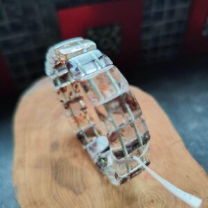 Puede incluir: Una pulsera de eslabones rectangulares transparentes con inclusiones de color marrón oscuro y marrón rojizo incrustadas en cada segmento. La pulsera está construida con un cordón interno flexible e incluye una pequeña etiqueta de plástico blanco.