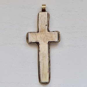 May include: A large, cross-shaped pendant with a cream-colored center and a gold-colored metal frame. The cross has a textured surface and a small loop at the top for a chain. The frame has a decorative, rope-like design.