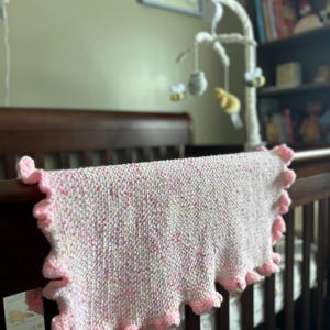 May include: A soft, hand-knitted baby blanket in a crib. The blanket is primarily white with pink and yellow flecks, and a ruffled pink border. The crib is dark brown, and the background includes a mobile and a framed picture.