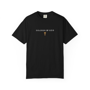 May include: Black t-shirt featuring the words "SOLDIER OF GOD" in white, with a gold cross beneath. The shirt has a crew neck and short sleeves. The design is centrally placed on the front.