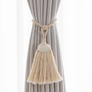 Può includere: Un fermatenda in corda beige con un design a conchiglia e nappa. Il fermatenda è avvolto attorno a una tenda grigia, creando un accento decorativo. La nappa è fatta di fibre morbide color crema.