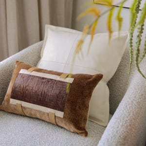 May include: A decorative pillow with a brown velvet cover and a rectangular, textured leather panel. The panel is secured with beige straps and white tape. A white pillow is behind it, resting on a gray upholstered chair.