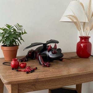 May include: A black and red dragon-like frog figurine with wings and horns, positioned next to a small red ceramic accessory with a black cord. The frog has a smooth finish with red accents on the eyes, horns, and feet, and black wings.