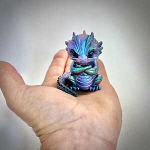 May include: A small, iridescent dragon figurine in shades of blue, purple, and green. The dragon has its arms crossed and a stern expression. The figurine is approximately 7.6 cm tall and has a detailed, textured surface.