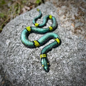 May include: A decorative snake figurine featuring a metallic green, scale-like textured body with distinct, bright yellow bands encircling its length. The snake has a pointed head with yellow eyes and is coiled in a serpentine shape on a surface.